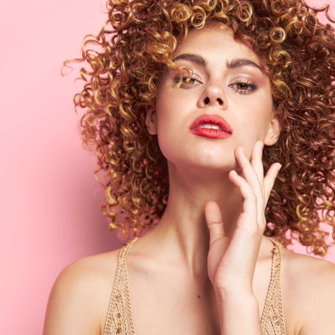 Woman with curly hair posing against pink background.