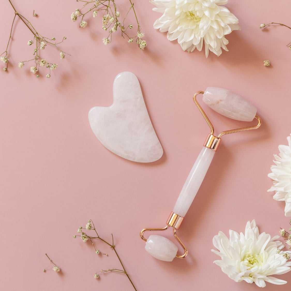 Jade roller and gua sha stone with white flowers on a pink background.